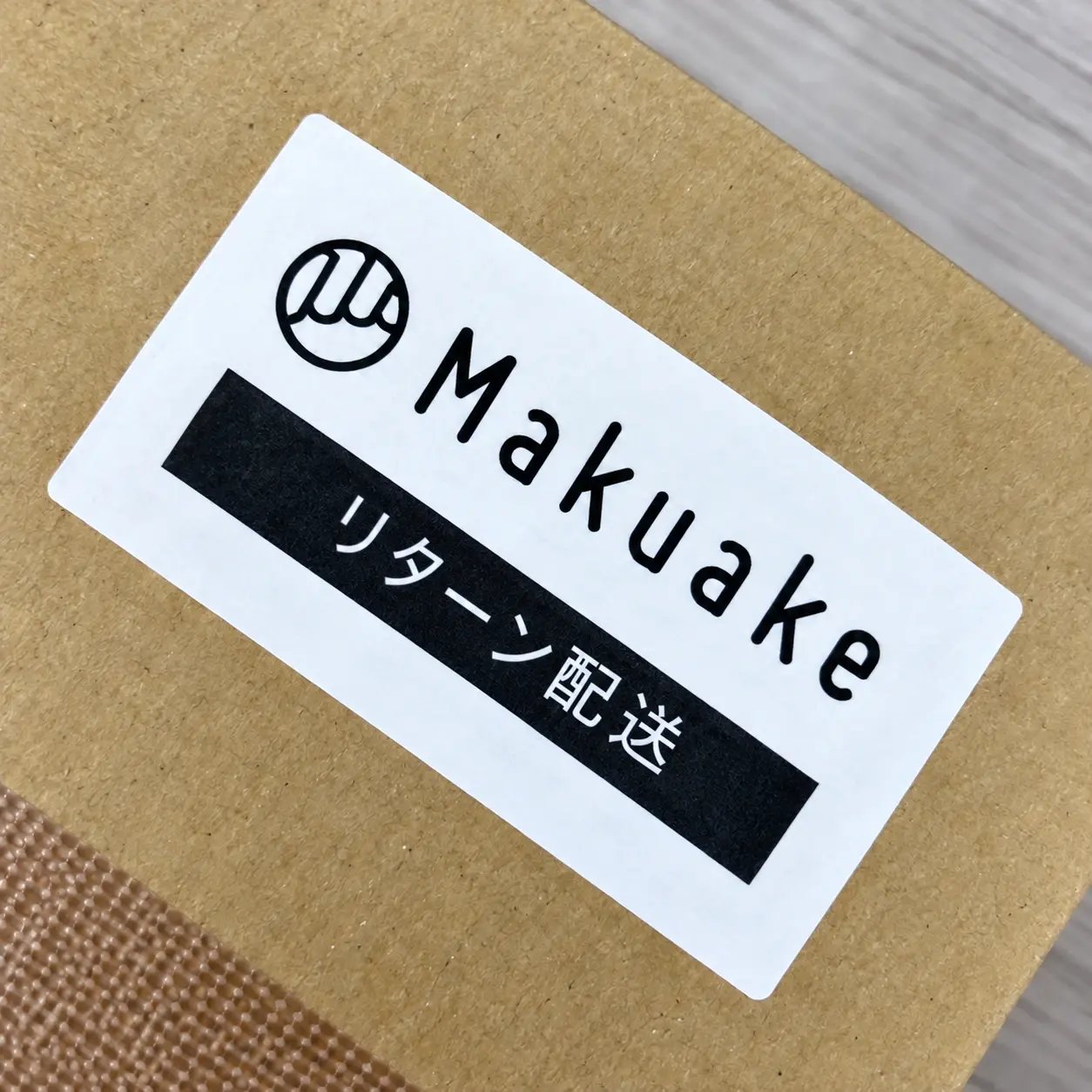 Makuakeのロゴと「リターン配送」の文字が入った専用シールを貼った段ボール